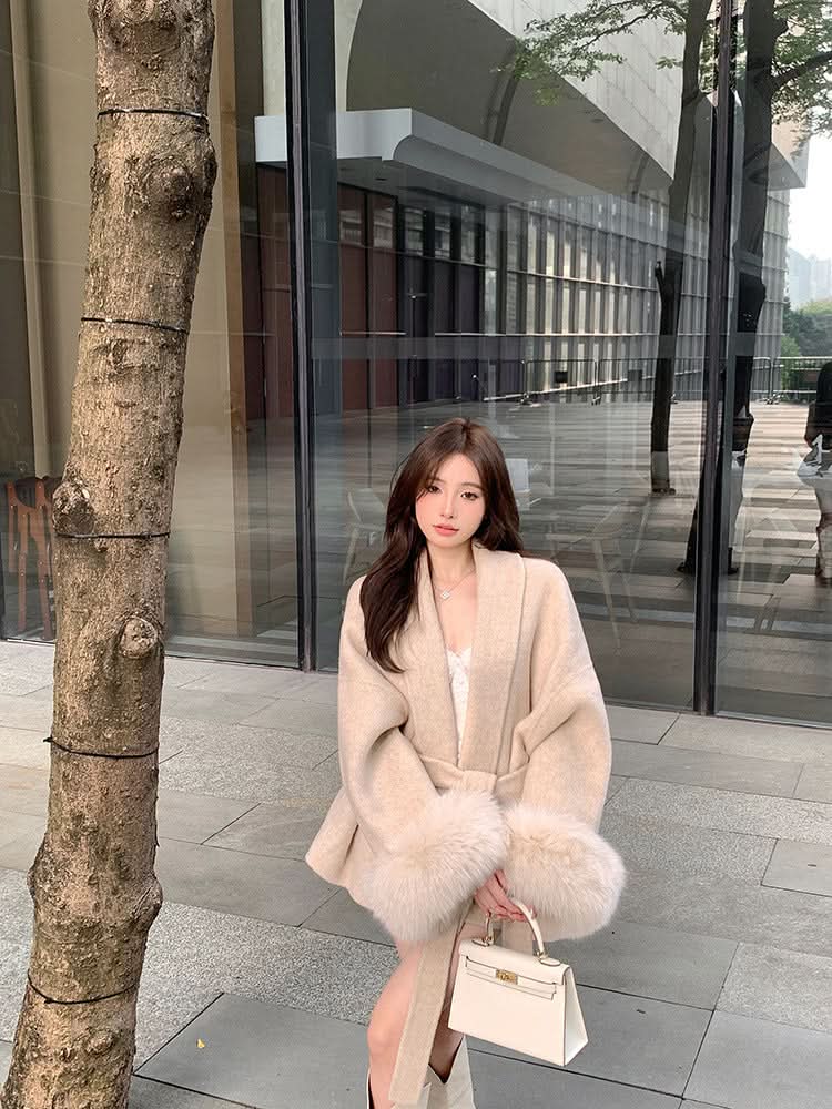 Beige wrap coat with fur cuffs worn with cream handbag in urban setting