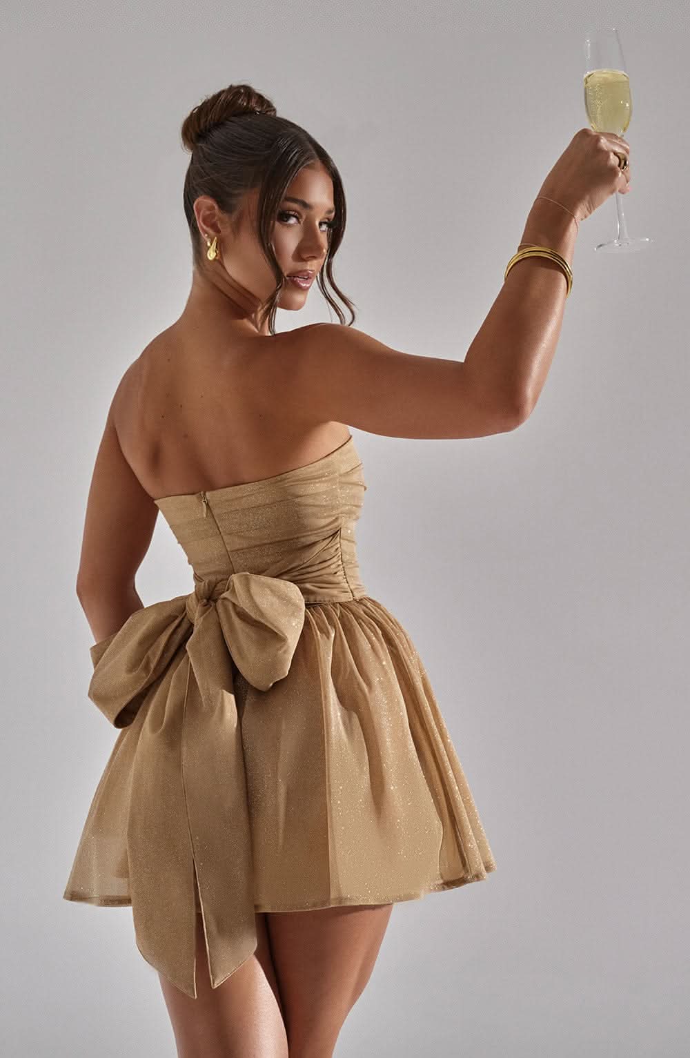 Back angle of gold sequin mini dress with large bow and champagne drink