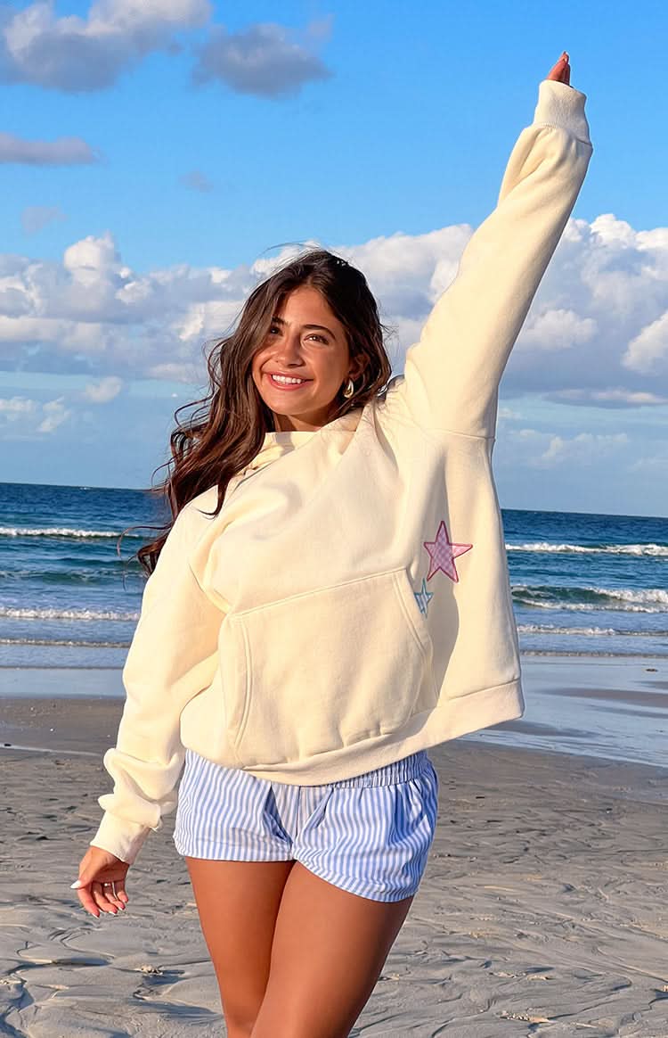 Cream oversized star hoodie with pink and blue star appliqués, worn on beach with arm raised